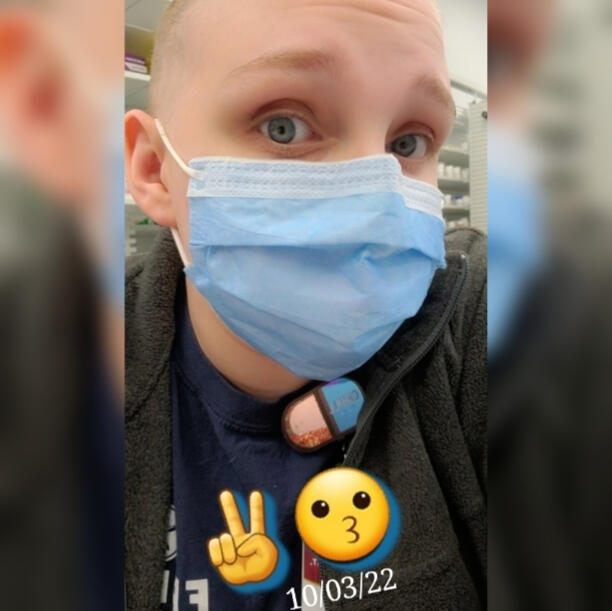 A selfie I took at work with me featuring my shaved head, me wearing a mask, & part of my name badge (a "chill pill" clip). Dated 10/3/22
